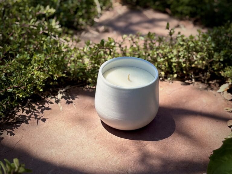 Organic candle in white ceramic vessel on red flagstone with greenery in the background.
