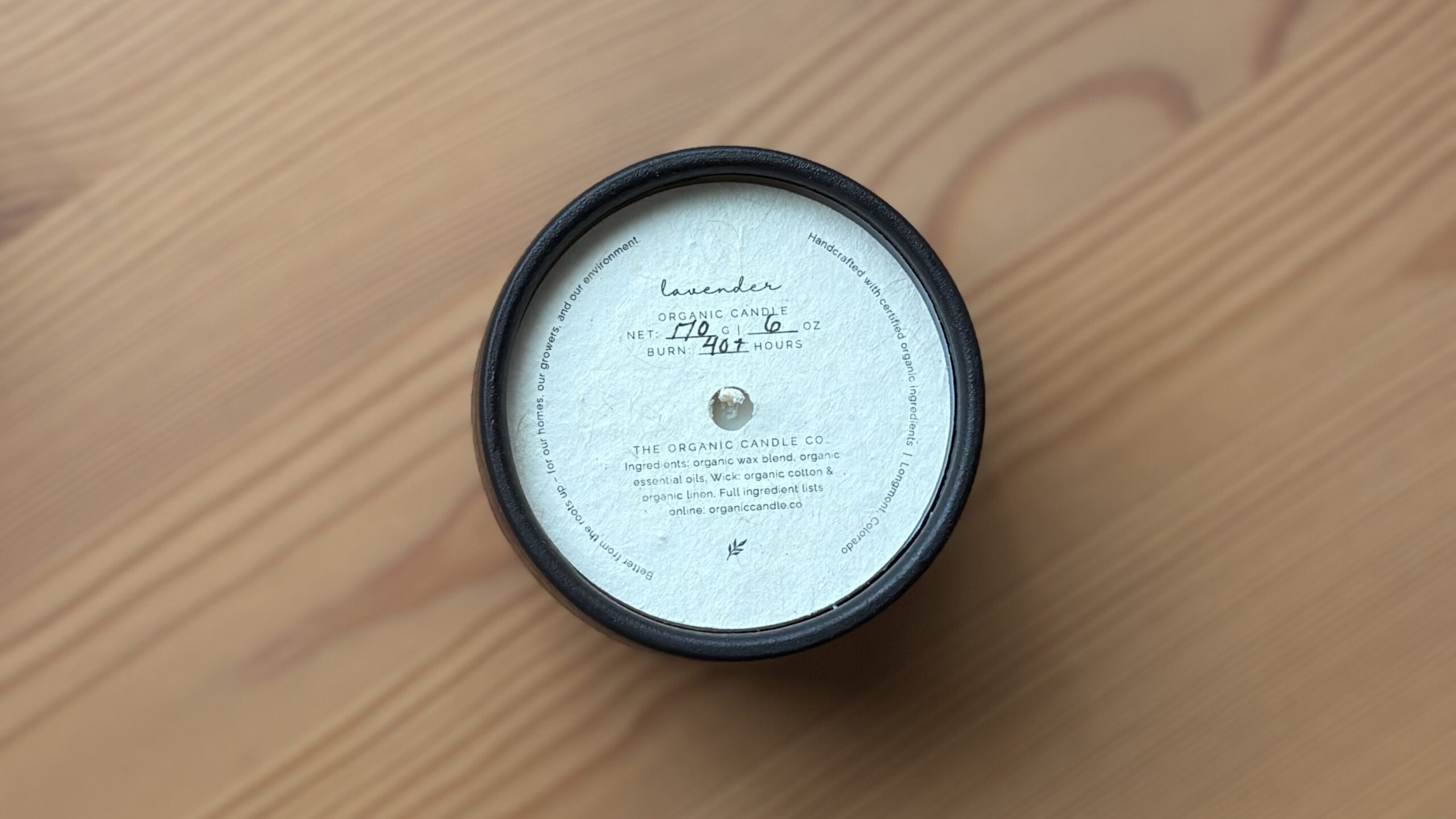 Organic candle in a black ceramic vessel with a dust cover on a wooden background.