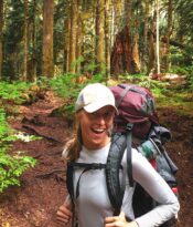Female wearing a hat and backpacking gear in the outdoors with trees in the background.